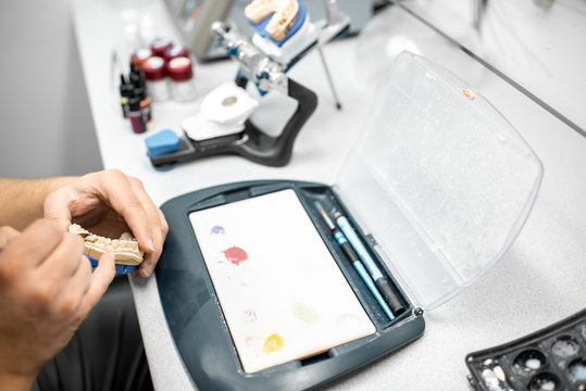 Dental technician coloring dental prosthesis with a paint brush at the laboratory, close-up view. Concept of implantats producing