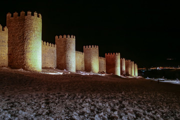 Muralla de Ávila iluminada en la noche y con un manto de nieve a sus pies.