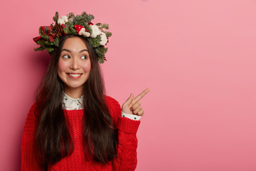 Beautiful cheerful woman with appealing look, points fore finger aside on blank space, smiles happily, shows advertising, dressed in warm outfit, Christmas wreath on head isolated over rosy background