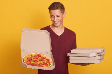 Handsome young delivery worker dresses burgundy casual t shirt holding pizza in boxes, looking at opened box and smiling, attractive male stands against yellow studio wall, looks happy, doing his job.