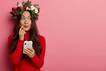 Photo of contemplative Korean woman uses modern cell phone for online communication and reading necessary information, wears decorative wreath, thinks deeply about something, isolated on pink