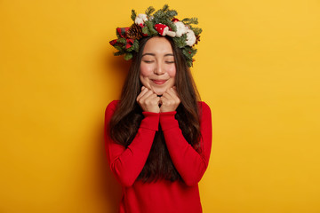 Adorable young lady has pleasant memories, smiles pleasantly, keeps hands under chin, wears festive wreath on head, recalls nice moment in mind, isolated over yellow background. Beauty, tenderness
