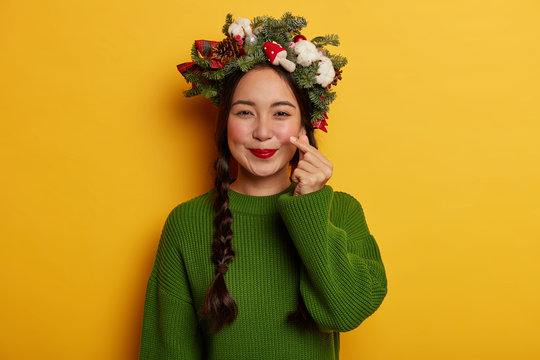 Attractive girl with dark pigtails, red painted lips shows sign of love, shapes heart with tip of thumb and index finger, expresses affection in korean style, wears beautiful Christmas wreath