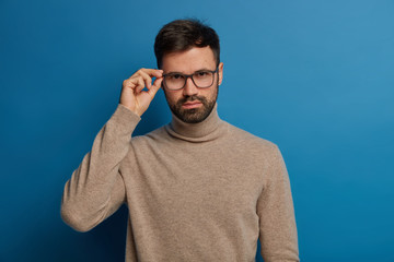 Handsome self confident man with thick bristle, keeps hand on rim of glasses, wears casual brown turtleneck, looks seriously at camera, has conversation with colleague, discuss their future plans