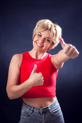 A portrait of young woman with short blond hair in red shirt showing thumb up symbol in front of black background. Lifestyle, emotions and beauty concept