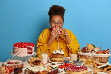 Fotobehang Funny glutton woman bites cakes with big appetite, cant stop eating sweet desserts, being in mood for enjoying sugary freshly baked bakery, surrounded by sweet treasures, has yummy snack, feels hunger © Wayhome Studio
