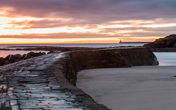 The stone pier at the harbour mouth of Cullercoats Bay on the coast of North Tyneside, England, UK. At sunrise with a colourful sky, taken at low tide.