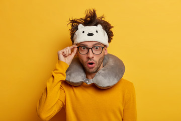 Terrified male passenger travels in bus, stares through optical spectacles, wears sleep mask and inflatable neck pillow for comfort travel. Shocked man plane tourist shocked by layover or stopover