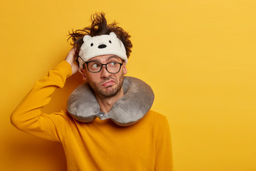Thoughtful contemplative man rubs head, thinks which seat in plane to choose, uses travel pillow to nap on long airplane flight, wears spectacles and eye flap, isolated over yellow wall, looks aside