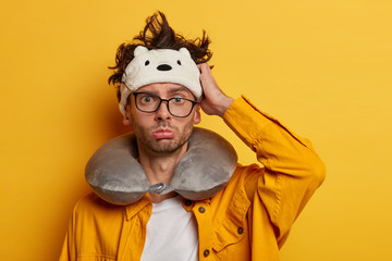 Tired Caucasian man has dissatisfied gloomy expression, travels by airplane or in car on long distance, awakes in bad mood during flight, wears transparent glasses, inflated neck pillow, sleep mask