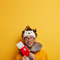 Smiling male tourist gets ready for plane flight, shows air tickets and passport, approcaches boarding pass, wears neck pillow for sleeping looks upwards with glad expression copy space on yellow wall