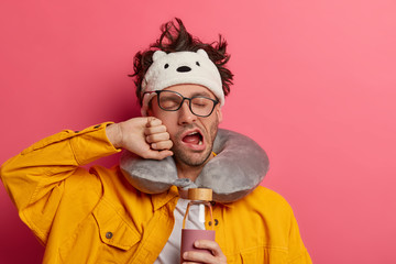 Photo of exhausted unshaven man traveler yawns from tiredness, wants sleep badly, holds glass...