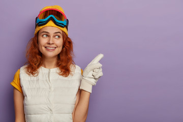 Positive redhead girl with gentle smile, wears yellow hat and white vest, points aside has happy mood after winter vacation, enjoys snowboading, poses indoor against purple wall. Happy snowboarder