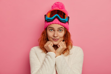 Portrait of active female snowboarder looks directly at camera with hands under chin, prepares for...