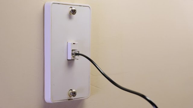 Male electrician plugging in landline telephone cord into wall mounted jack plate.