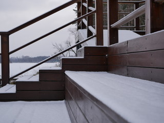 The brown wooden staircase is covered with a layer of snow without traces. Steps with railings to the river on a cloudy winter morning.