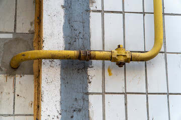 Little yellow gas crane on the pipeline near wall with white tiles. Pipe with crane allows to distribute natural gas and turn it off if necessary. On the conduit located pipe coupling and elbows