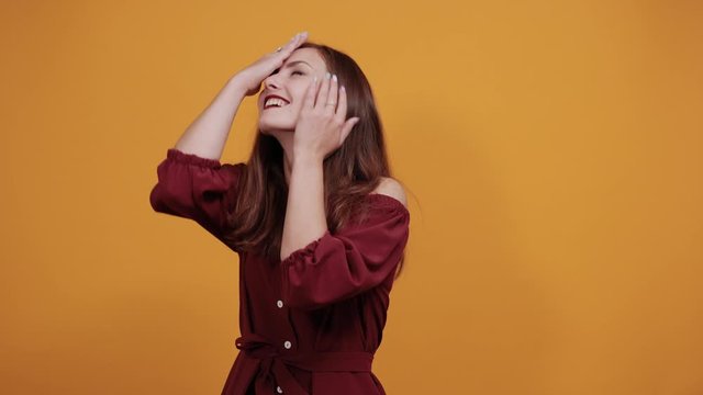 Cheerful charming lady in fashion maroon dress looking directly, spreads hands, smiling isolated on orange background in studio. People sincere emotions, lifestyle concept.