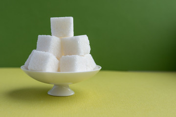 Cube sugar in a white bowl on a leg on a green background. Close-up on a colored natural...