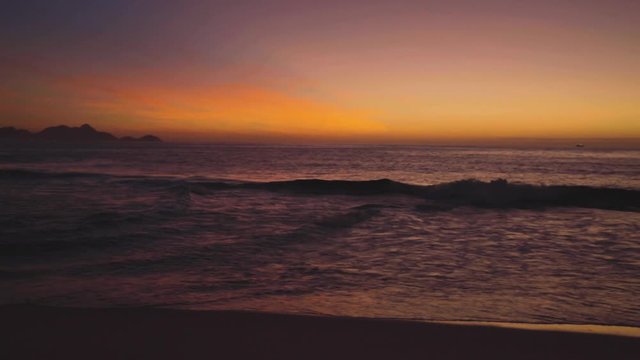 Pan movement around the pristine Devils beach in Ipanema, Rio de Janeiro, at daybreak with waves coming in and a dark orange glow from the sunrise on the horizon and water in front