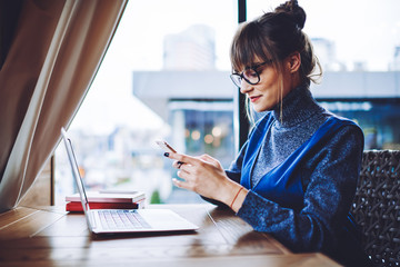 Millennial hipster girl in spectacles checking notification on modern cellular phone during break from e learning on laptop computer, caucasian female freelancer typing text message to colleague
