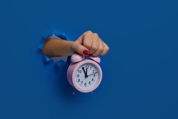 Wake up. Pink alarm clock in hand through a torn dark blue paper wall, mockup. Space for text. Run...