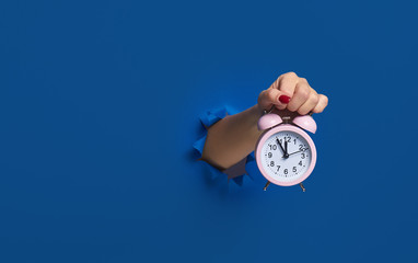 Wake up. Pink alarm clock in hand through a torn dark blue paper wall, mockup. Space for text. Run...