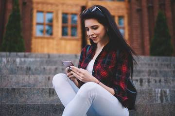 Positive smiling hipster girl sitting at street and using mobile phone for checking mail,happy cheerful woman installing application and reading message on modern cellular phone while resting outdoors