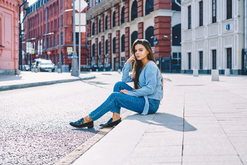 Fototapeta premium Portrait of young hipster girl sitting for little rest while spending time on city street, attractive female student in fashion apparel looking at camera and recreating outdoors on urban setting