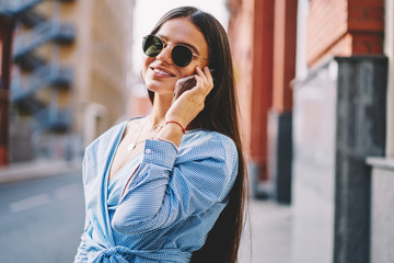 Fototapeta premium Charming and smiling hipster girl communicating over mobile phone with best friend outdoors, attractive woman in sunglasses and blue shirt calling using modern device and roaming connection