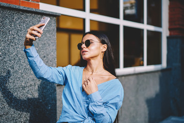Attractive stylish girl in stylish sunglasses taking selfie photo while walking at street, trendy hipster female blogger 20 years old in blue shirt making new video vlog using modern cellphone