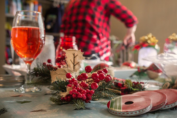 Christmas craft filled kitchen table with holly berries and rose wine. Women with red plaid shirt in background