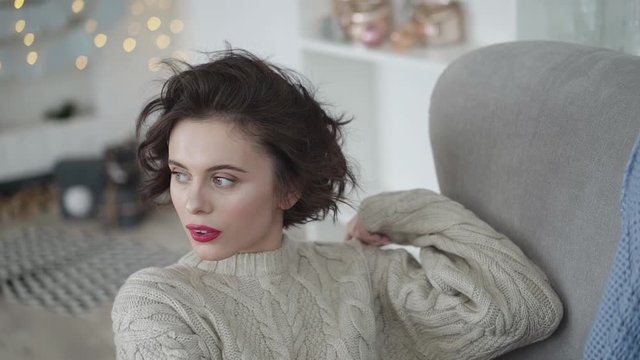 Glamorous brunette in warm sweater lying on armchair and smiling at camera