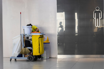 Female worker preparing janitorial car for cleaning in front of male toilets.