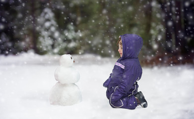 Curious little girl in purple snowsuit kneeling in snow looking at small snowman