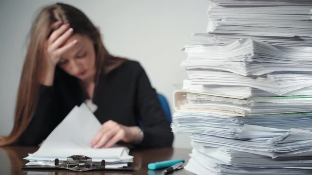 A pile of unfinished documents and files laying on desk in the office. Overworked female office accountant sitting in front of paperworks and holding her head with hands, tired worker having headache
