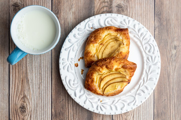 Fresh individual apple tarts on a white plate, blue cup of milk, wood background