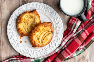 Fresh individual apple tarts on a white plate, cup of milk, red, green, and white cloth napkin, wood background