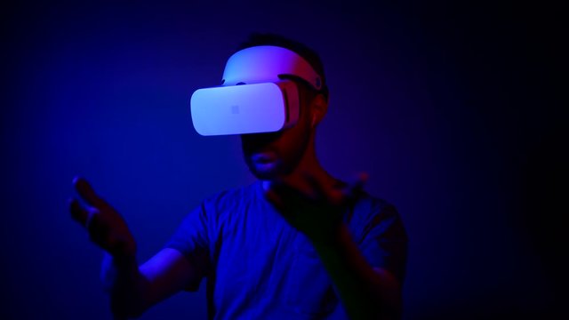 A man with a beard in a VR helmet interacts with virtual reality and views media content. A new futuristic way of interacting with technology. In trendy neon colors blue and red.