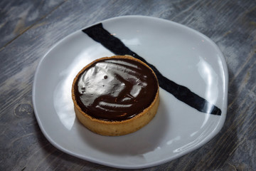 Tartelette au chocolat, a typical French pastry chocolate tart pie, on display on a white plate on a rustic wooden table. It is typical from the French patisserie cuisine