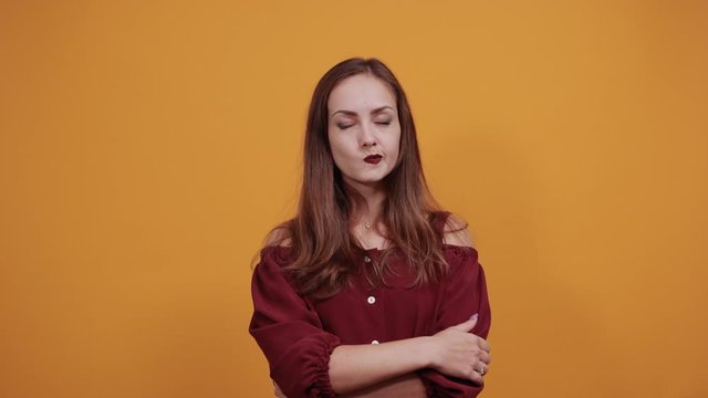 Focused caucasian woman in maroon dress isolated on orange background in studio keeping finger on cheek, squeezing mouth, thinking. People sincere emotions, lifestyle concept.