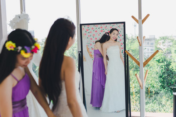 The bridesmaid is helping the beautiful bride wearing a white wedding dress in front of the mirror.
