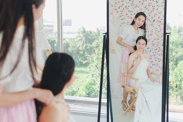 The bridesmaid is helping the beautiful bride wearing a white wedding dress in front of the mirror.