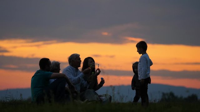 Children, parents and grandparents silhouettes play with bubbles on sunset hill,three different generations spend wonderful time together in nature, strong family relationship