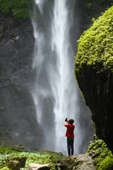 Obraz premium Stunning view of a tourist taking photos with a smartphone at the Tumpak Sewu Waterfalls. Tumpak Sewu Waterfalls also known as Coban Sewu are a tourist attraction in East Java, Indonesia.