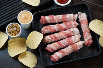 Cast-iron pan with raw fresh bacon wrapped sausages, high angle view over dark brown metal surface