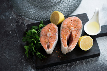 Fresh uncooked salmon steaks with parsley and lemon on a black wooden cutting board, studio shot over dark-grey stone background