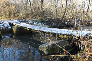 A small wooden bridge is covered in snow. On the street, in autumn, in some places on the earth there is snow and thaws are visible. Reflections of autumn trees are visible in the water of the river
