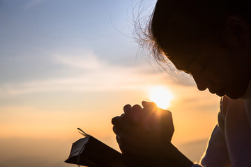 Silhouette of christian young woman praying with a  cross and open the bible at sunrise, Christian...