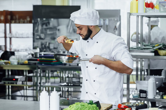 Happy successful young chef in white uniform preparing a meal with various vegetables.